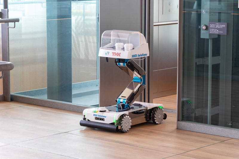 Lifter付きSEED-Mover。開発元はTHK。軽食や飲料の運搬を想定したパーソナルサイズの小型配膳ロボット。この日は2階の通路から3階のミーティングスペースの前までホットコーヒーを運搬。フロア移動に使うエレベータは自動で乗り降りする（エレベータの操作は手動）