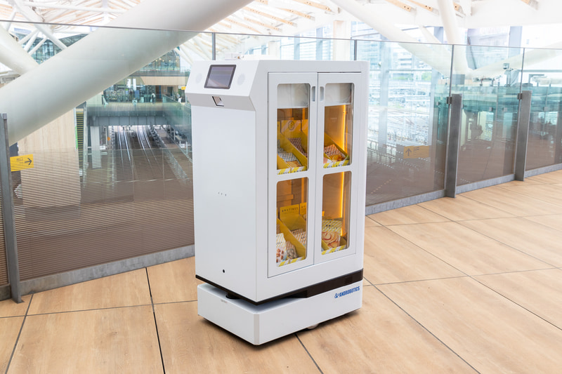 FRUTERA（フルテラ）。開発元はアンドロボティクス。軽食や飲料を目的地まで運ぶ搬送ロボットで、通信障害に強いWi-SUN無線通信を採用しているのが特徴。ミーティングスペースに準備された操作ボタンを押すと、通路に待機していた本機がスペース内のテーブル近くまでお菓子を運搬。終了操作をすると自動的に転回して元の位置まで戻った