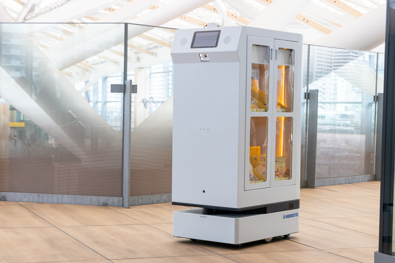 FRUTERA（フルテラ）。開発元はアンドロボティクス。軽食や飲料を目的地まで運ぶ搬送ロボットで、通信障害に強いWi-SUN無線通信を採用しているのが特徴。ミーティングスペースに準備された操作ボタンを押すと、通路に待機していた本機がスペース内のテーブル近くまでお菓子を運搬。終了操作をすると自動的に転回して元の位置まで戻った