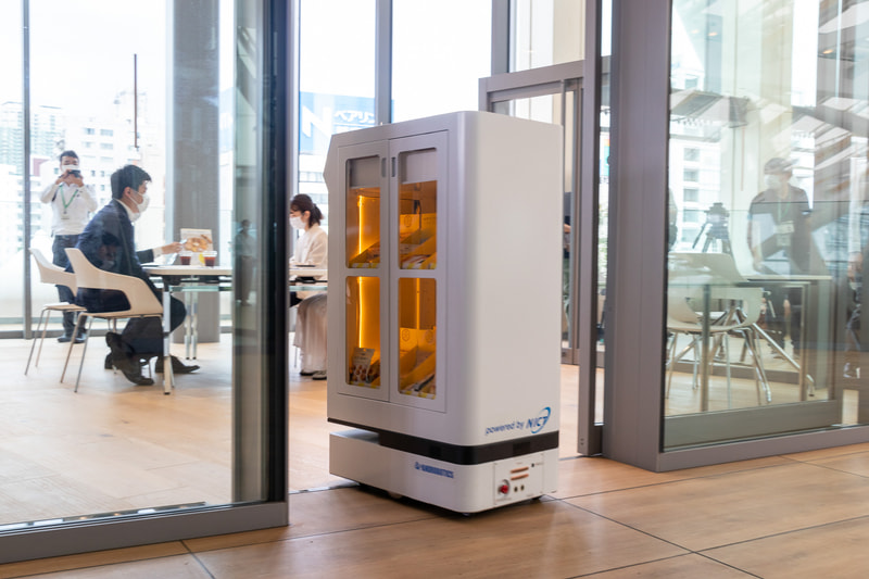 FRUTERA（フルテラ）。開発元はアンドロボティクス。軽食や飲料を目的地まで運ぶ搬送ロボットで、通信障害に強いWi-SUN無線通信を採用しているのが特徴。ミーティングスペースに準備された操作ボタンを押すと、通路に待機していた本機がスペース内のテーブル近くまでお菓子を運搬。終了操作をすると自動的に転回して元の位置まで戻った