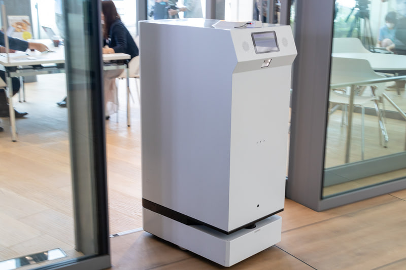 FRUTERA（フルテラ）。開発元はアンドロボティクス。軽食や飲料を目的地まで運ぶ搬送ロボットで、通信障害に強いWi-SUN無線通信を採用しているのが特徴。ミーティングスペースに準備された操作ボタンを押すと、通路に待機していた本機がスペース内のテーブル近くまでお菓子を運搬。終了操作をすると自動的に転回して元の位置まで戻った