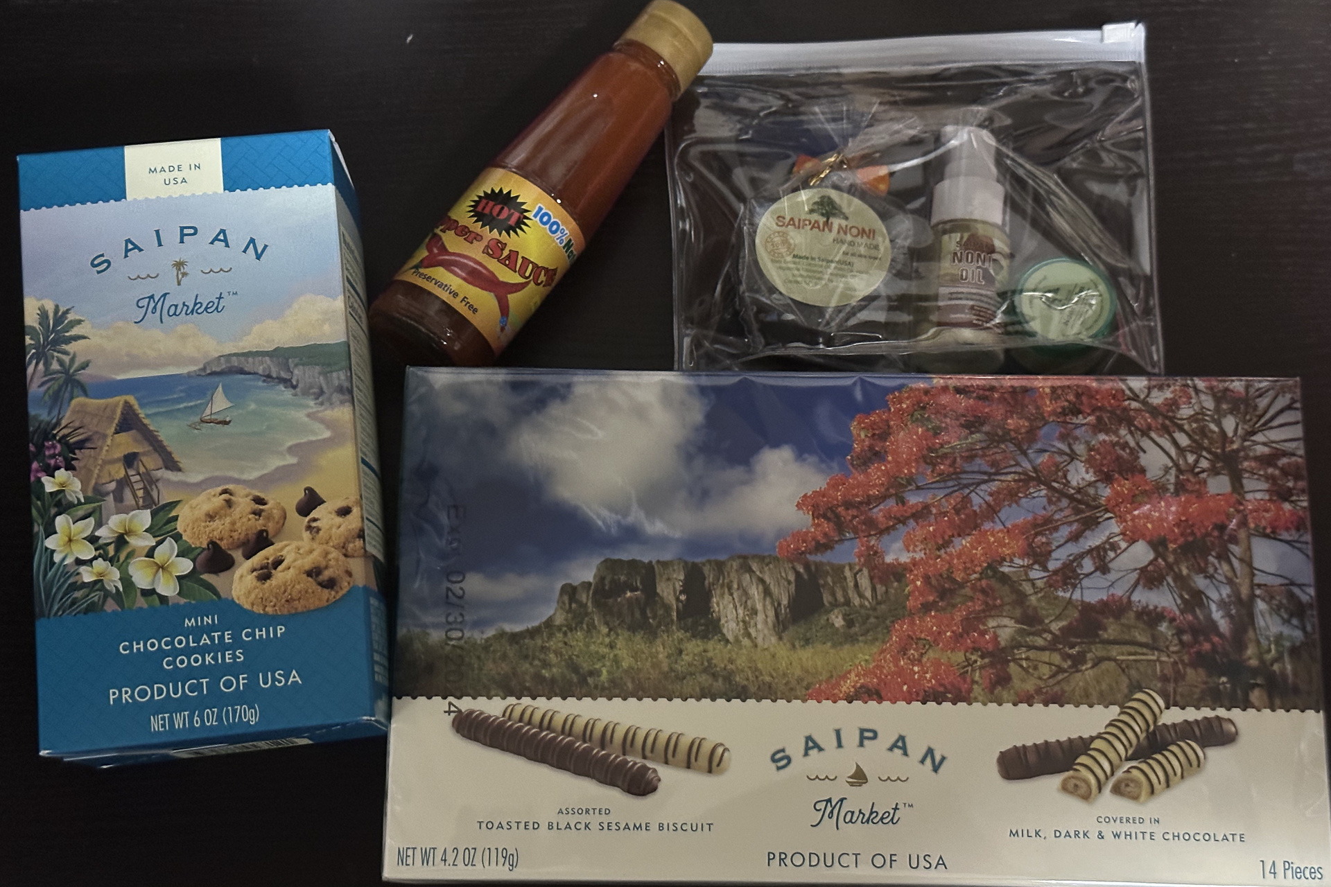 旅の醍醐味、サイパンで購入したお土産の一部。太平洋のアメリカでよく見かける風なチョコレートやクッキーなどはここでも。真っ赤なソースの瓶はテニアン島のチリペッパーを使ったホットソース（時期によってはないこともあるがフィナデニソースも売っている）。その右は熱帯ハーブの一種、ノニを用いた石鹸やバーム、オイルのセット。ここにはないがノニ茶も人気