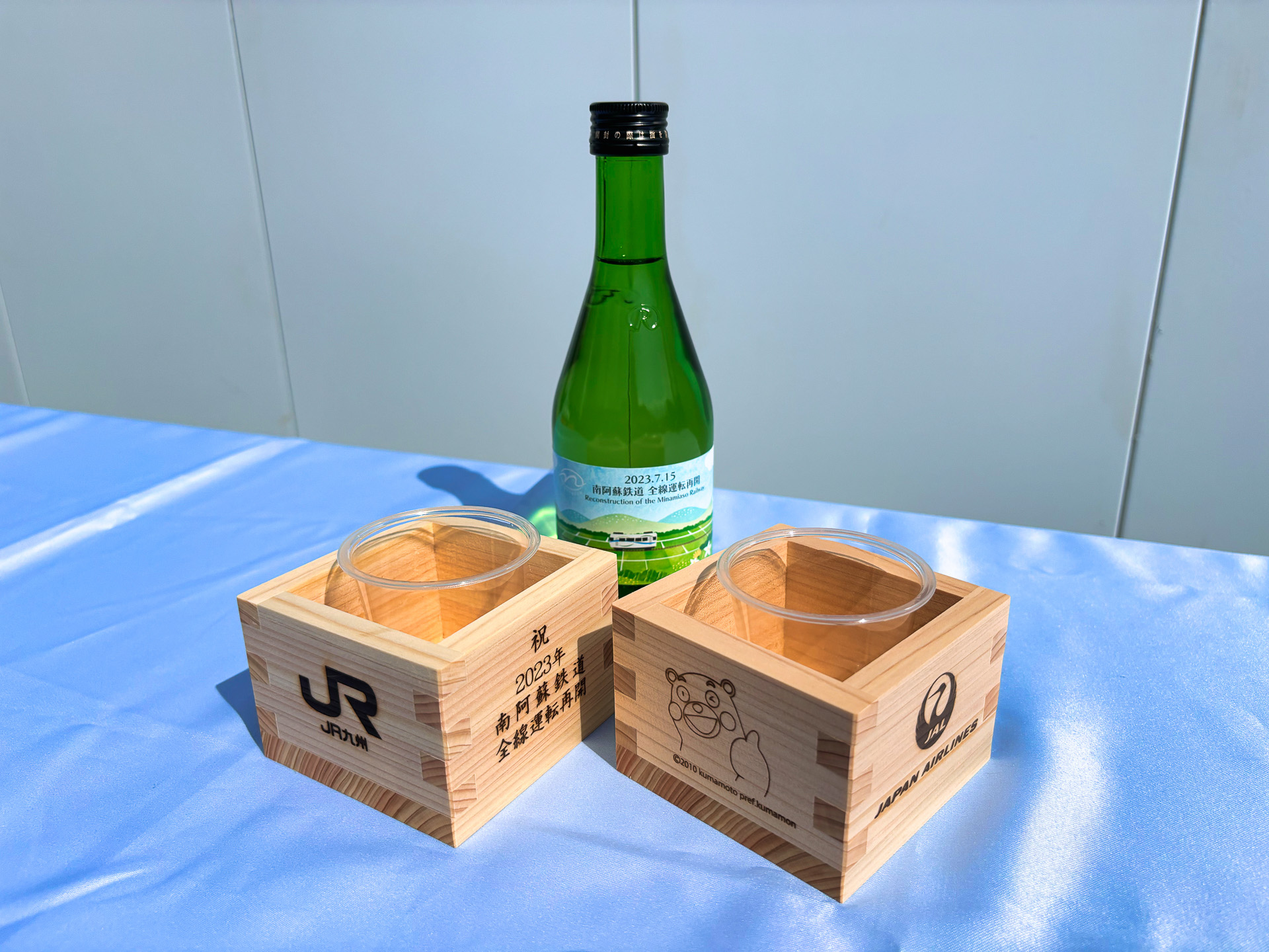 JR九州とJALとの共同企画で実施されたツアー。南阿蘇鉄道全線運転再開の幟が爽やかな風になびいていた。記念の枡と阿蘇の酒 れいざんの特別ボトルが配布された