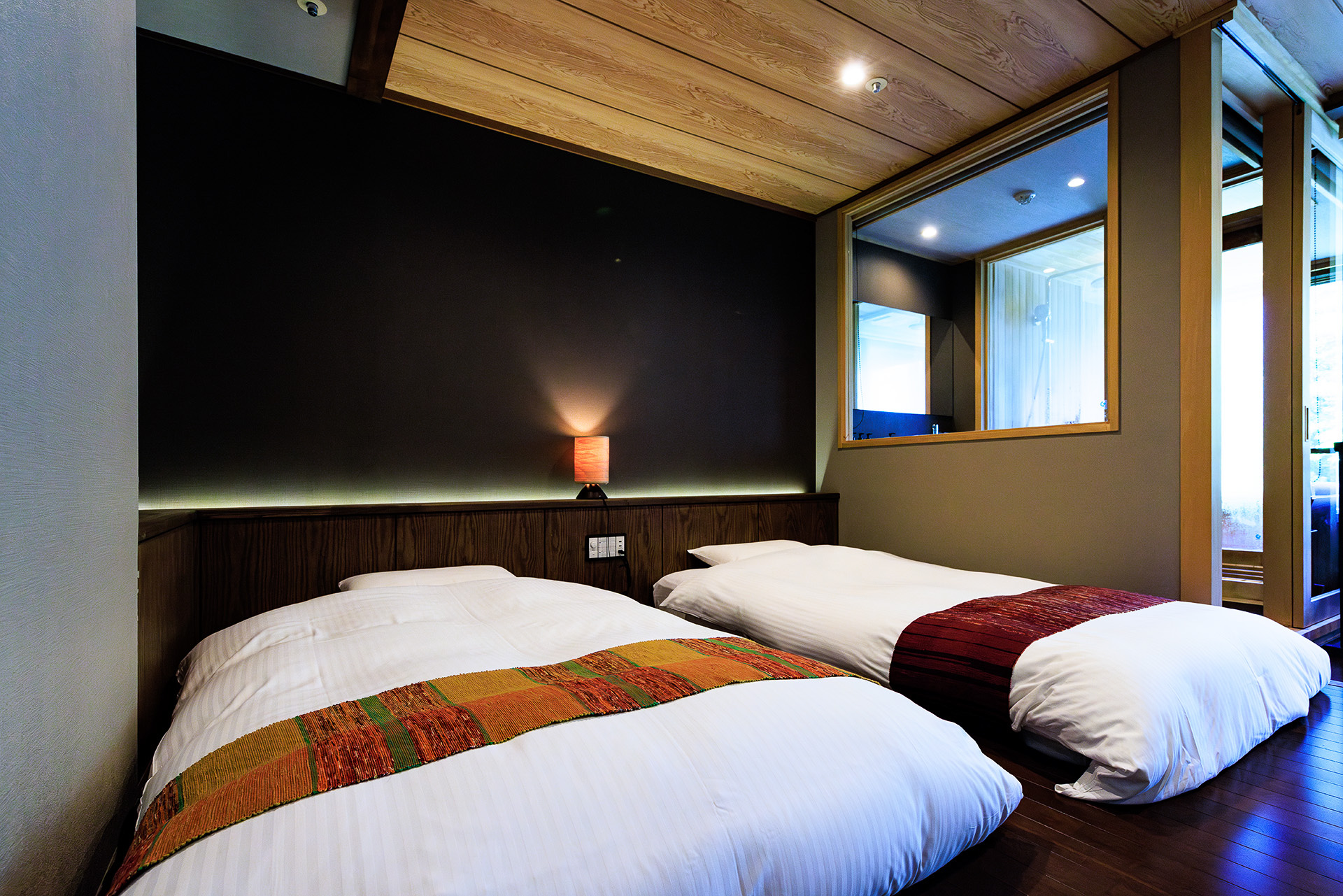 畳の床より1段高いところにローベッドがある。ベッドスローは「南部裂織（なんぶさきおり）」、枕元には青森のブナの木を利用したBUNACO製のテーブルランプも