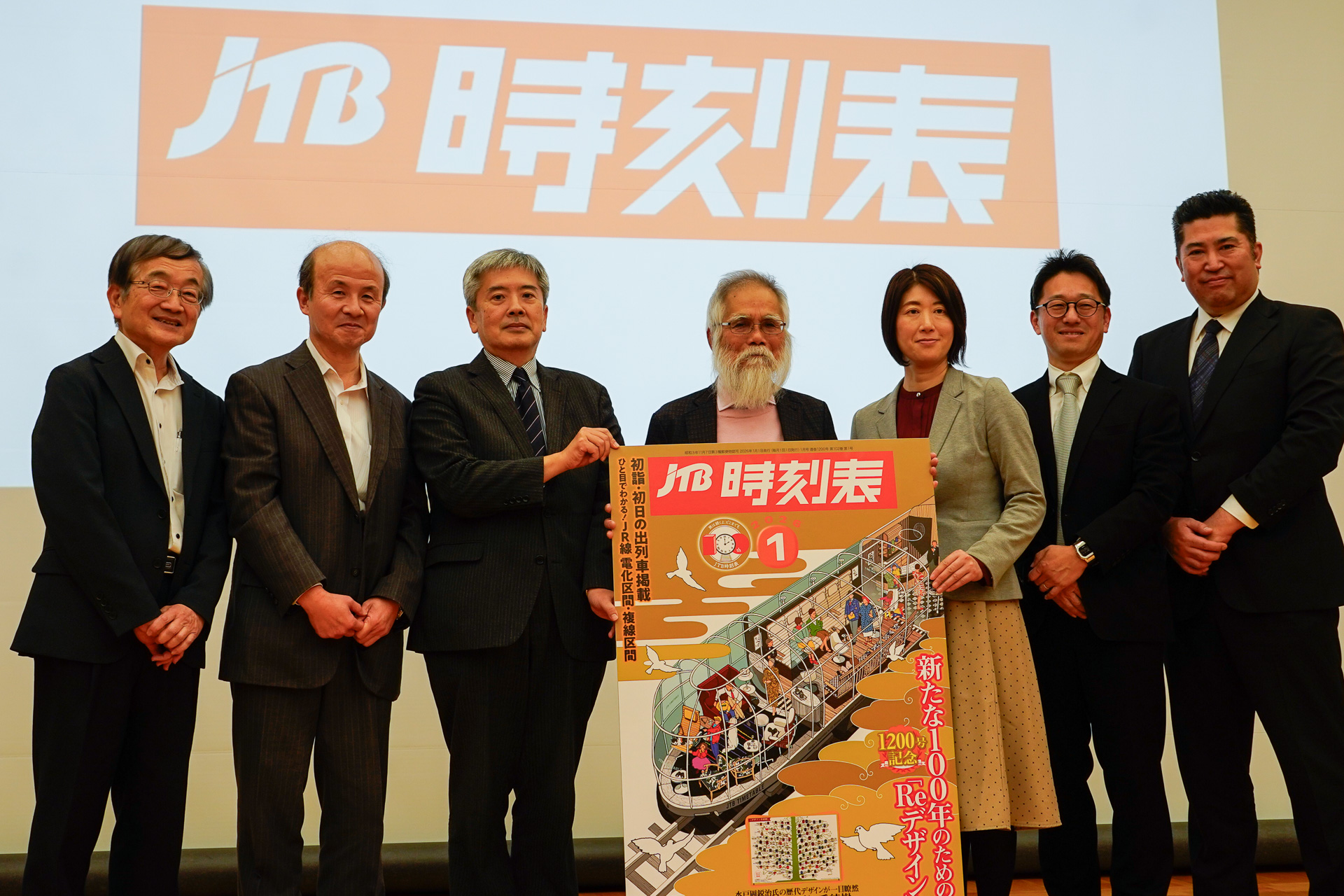「JTB時刻表」の新ロゴをバックに、1200号の表紙パネルを持って並ぶ歴代編集長。左から順に、木村嘉男氏（第11・13代編集長）、高山法悦氏（第14代編集長）、石川敏晴氏（第16代編集長）、石野哲氏（第8代編集長）、梶原美礼氏（第19代編集長）、田代浩一氏（第18代編集長）、大内学氏（第17代編集長）