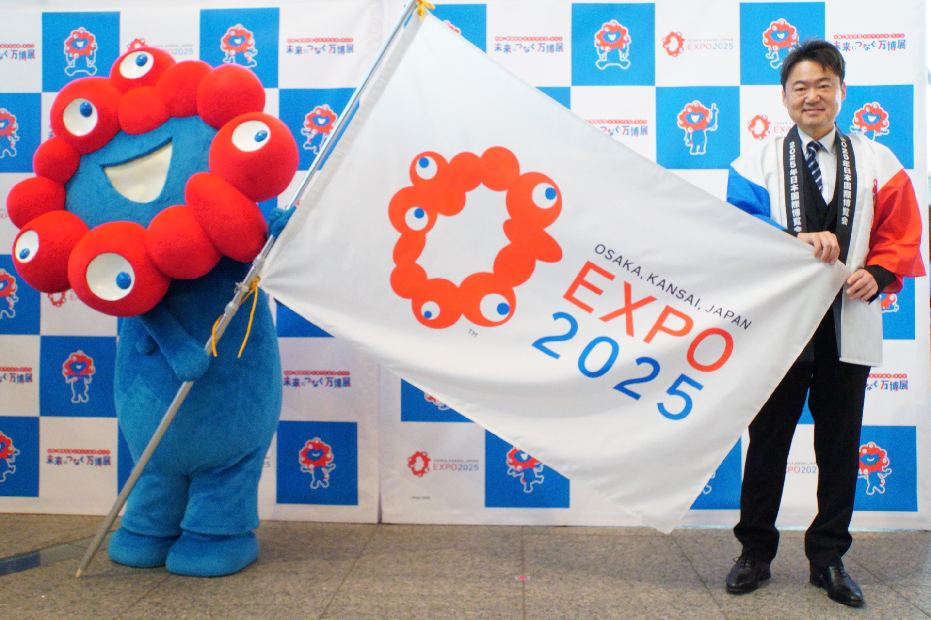 内閣官房 内閣審議官 国際博覧会推進本部 事務局次長 井上学氏からミャクミャクへ、メモリアルフラッグの受け渡しが行なわれた