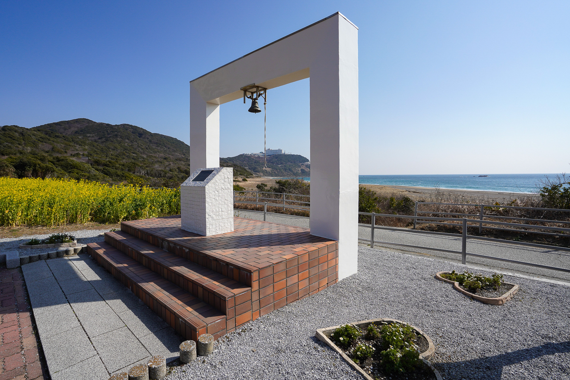 菜の花と海に囲まれて、真っ白な鐘の記念碑が建てられている