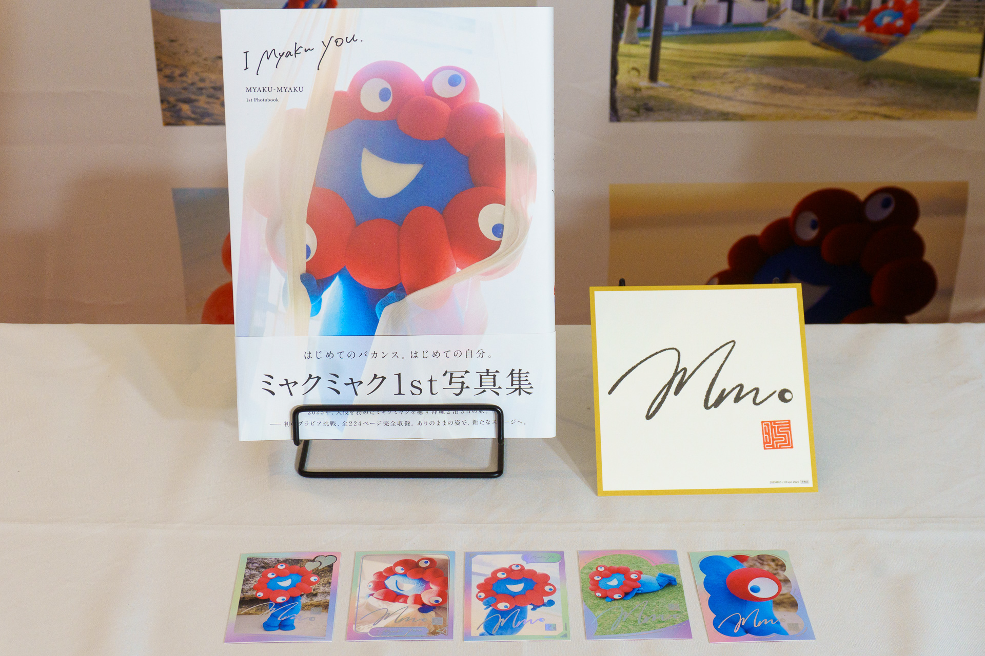 奥に並んでいるのは、1stグラビア写真集「I myaku you.」（左）、ミャクミャクによる落款印が押されたサイン色紙（右・お渡し会限定特典）。手前はランダム1枚で封入される特製フォトカード（全5種類）