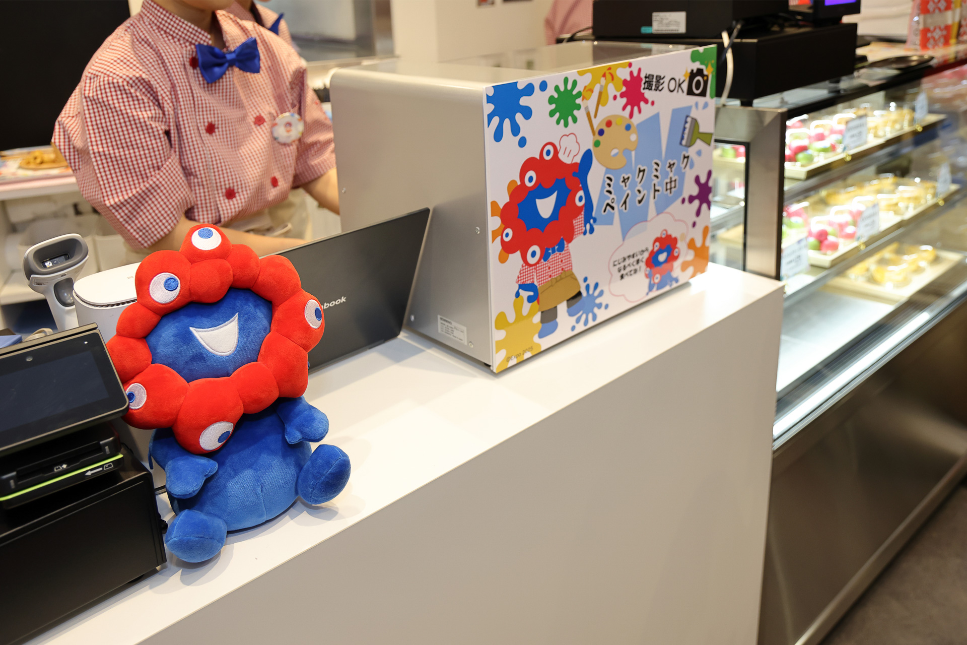 レジ（開店準備中）のまわりにもミャクミャク。チーズケーキの絵柄はプリンターで描かれる。撮影OK