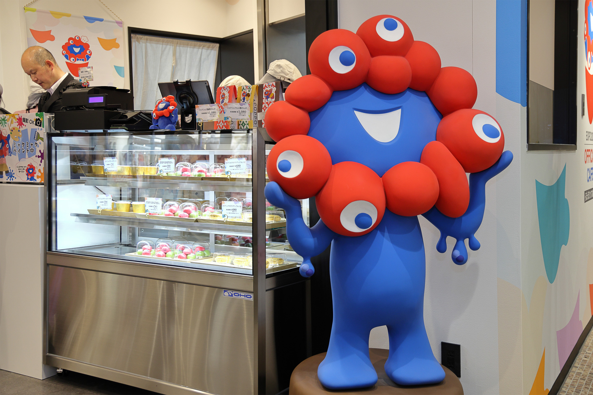 レジ（開店準備中）のまわりにもミャクミャク。チーズケーキの絵柄はプリンターで描かれる。撮影OK