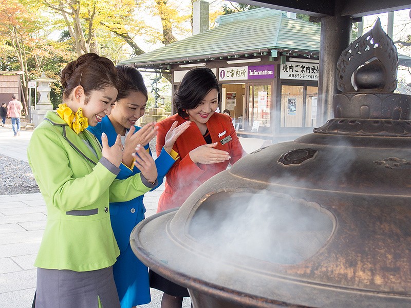 常香炉で身を清めてから参拝に向かうCAの3人。左からSpringJapan（春秋航空日本）の和田朱里さん、バニラエアの杉山友梨さん、ジェットスター・ジャパンの塩満萌加さん。説明をするのは成田山新勝寺の松岡照英氏